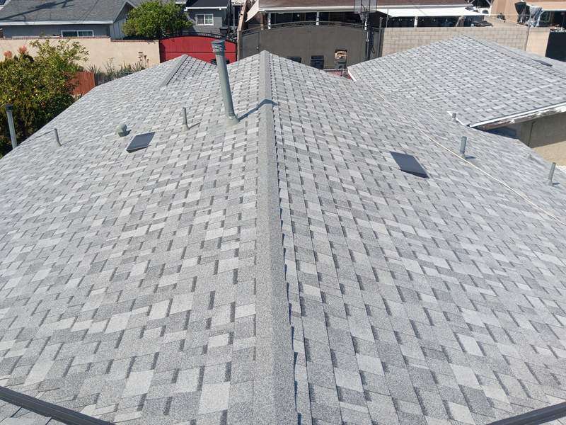 Gray asphalt shingle roof planes meeting at hips and a ridge, with several box vents and surrounding residential roof context under daylight.
