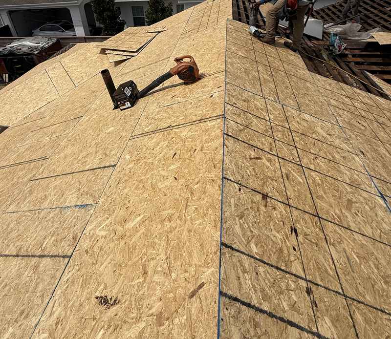 A bare tan OSB roof deck over a hip roof with panel seams and nail lines clearly visible, a pneumatic nailer and its hose lying on the deck, and a small stack of material near the ridge under a clear blue sky.