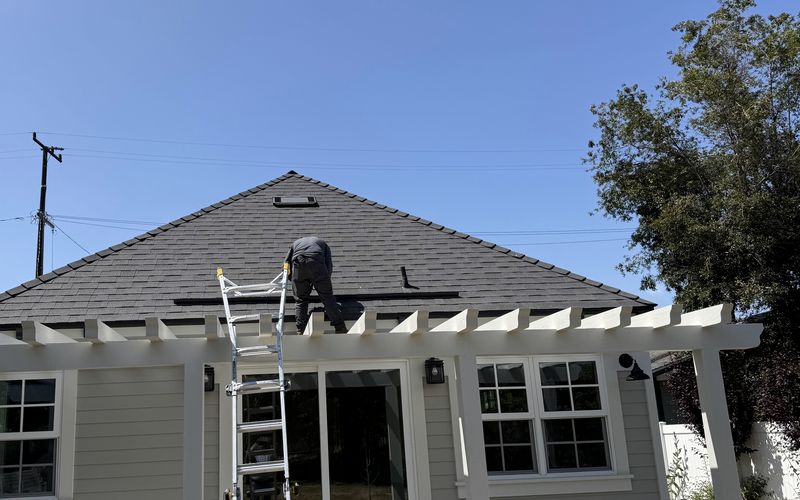 A worker on a multi-position aluminum ladder reaching the eave of a gray asphalt shingle roof on a single-story house, with a pergola-covered patio, chairs and garden foliage below and a clear sky above.