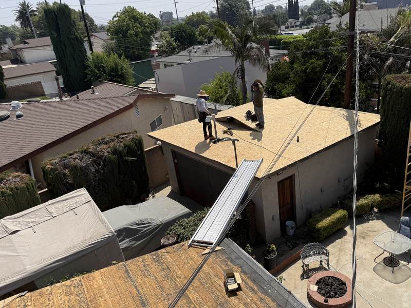Broad wood roof deck with marked panel layout, a worker, staged materials, a ladder, and surrounding residential job context.