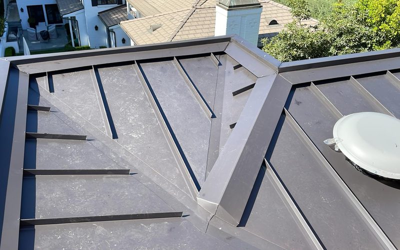 Standing seam metal roof panels meeting at a hip ridge on a residential home, protective film still on the pans and a round vent cap on the adjacent slope