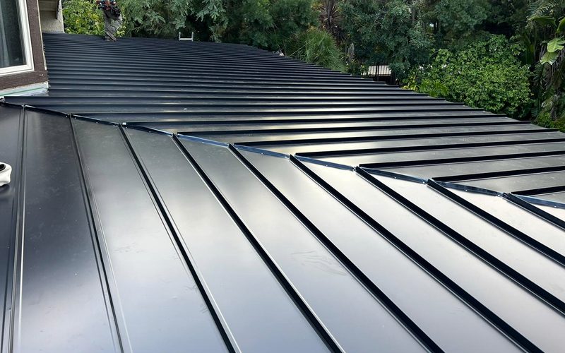 Dark standing-seam metal roof planes meeting at a stepped ridge, with continuous raised seams across the plane and mature landscaping behind.