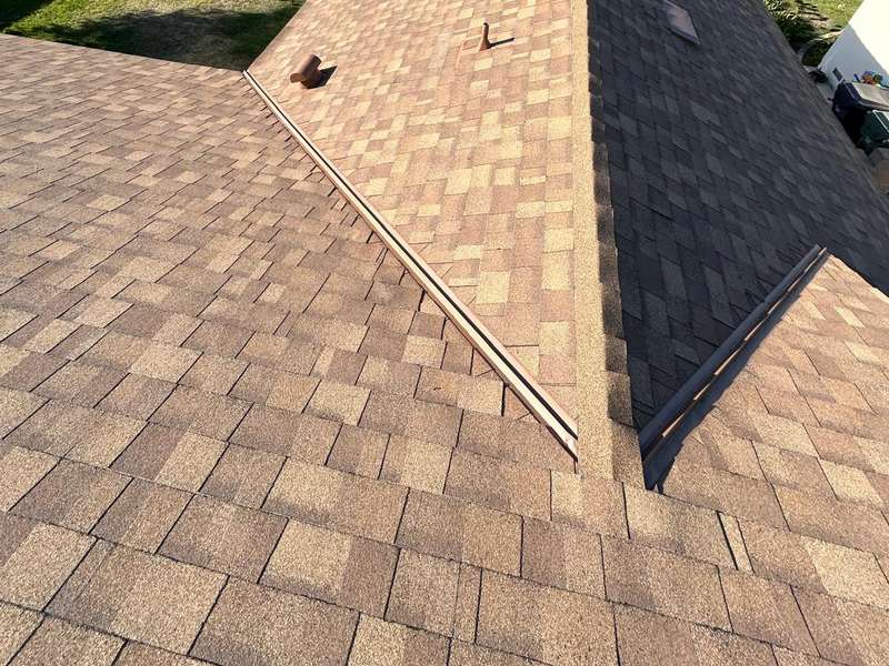 Elevated view of a tan and brown asphalt shingle roof with hip ridges and a valley, a dark charcoal shingle plane adjoining at right, a vent boot near the ridge, and a wooden plank lying across the shingles, with trees beyond the roof edge.