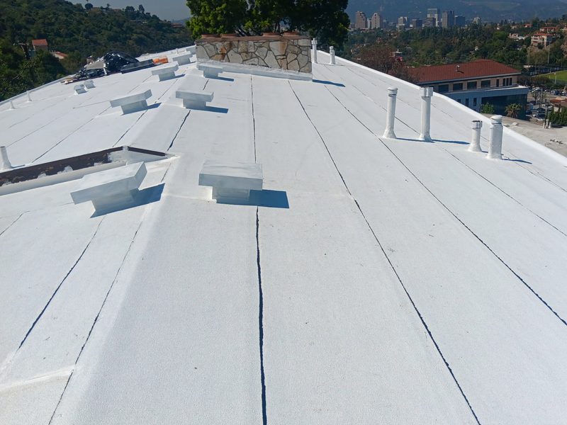 Rooftop-level view of a finished white-coated low-slope roof running to a crown, with long seam lines, six vent stacks, several metal curb caps, a stone-faced chimney and a distant mountain and skyline horizon.